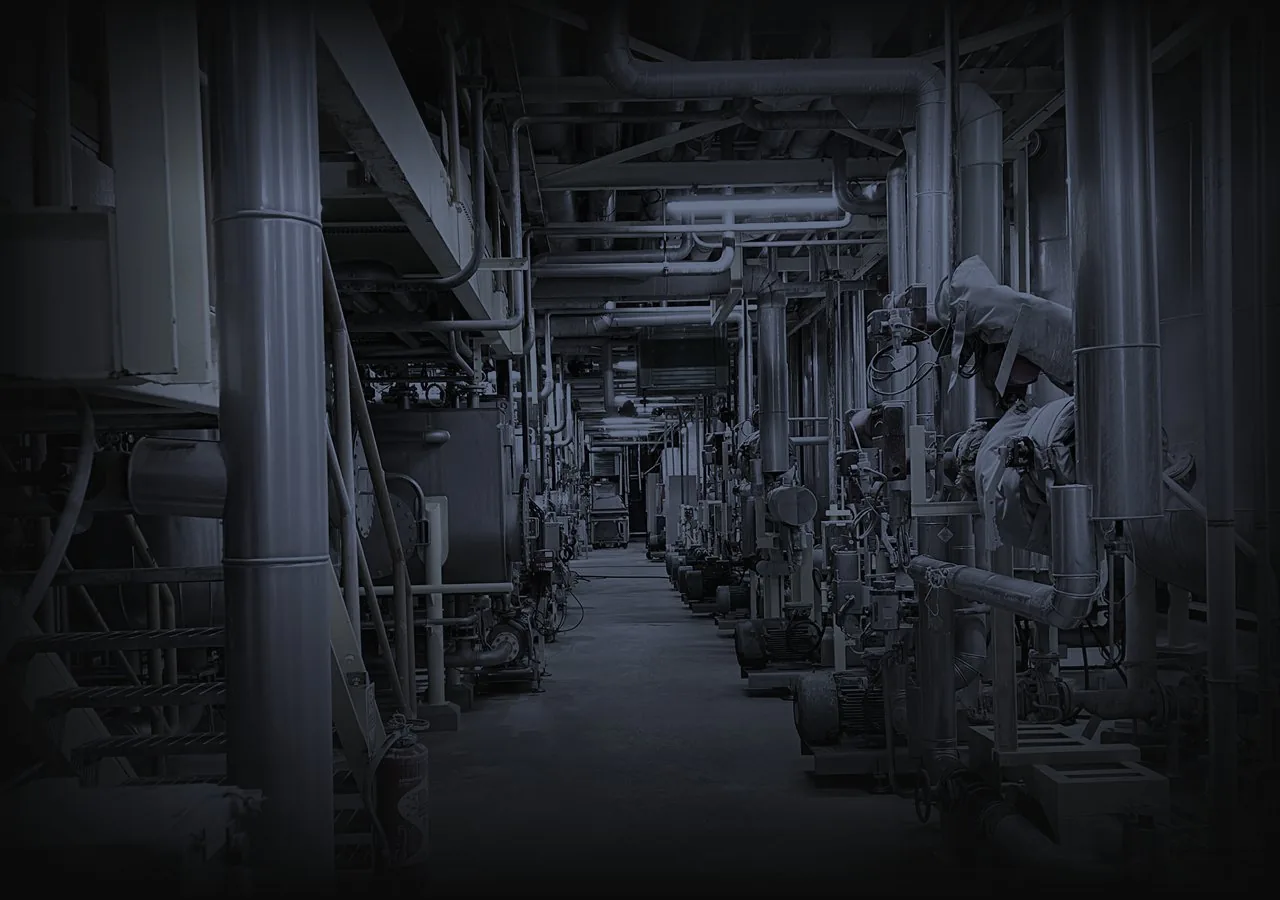 Allee technique d'une usine : tuyauteries, pompes et moteurs alignes de part et d'autre, sous un eclairage au neon. Personne.